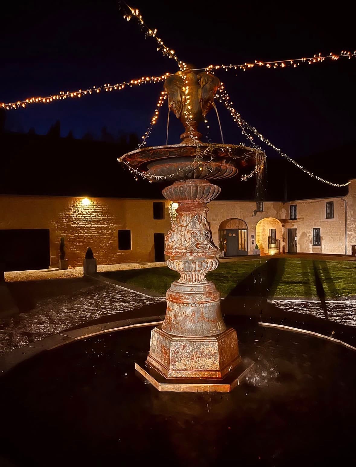 Fontaine d'époque en fonte de 4 mètres au centre de la Cour d'Honneur du Domaine de la Cour des Lys, illuminée la nuit sous des guirlandes lumineuses