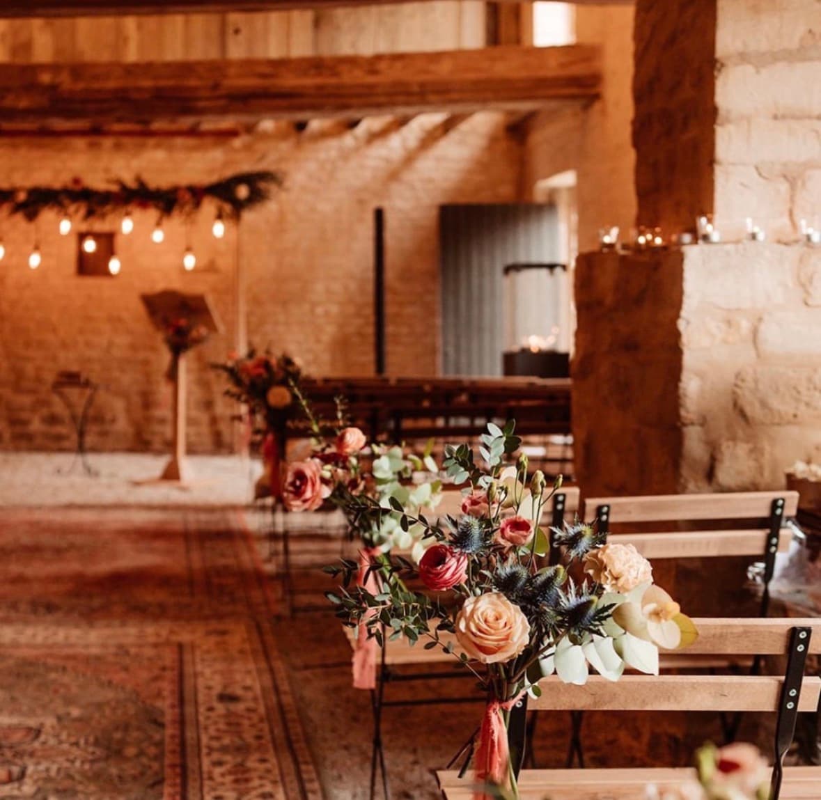 Préau couvert du Domaine de la Cour des Lys, dressé pour une cérémonie laïque avec bancs en bois, bouquets de fleurs, guirlandes lumineuses et tapis ancien